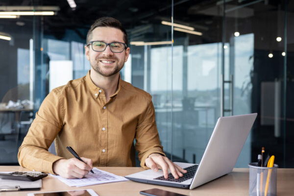 Portrait of young businessman in shirt, man smiling and looking at camera at workplace inside office, accountant with calculator behind paper work signing contracts and financial reports.