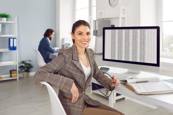 Portrait of professional financial accountant at work. Happy cheerful beautiful young woman in suit sitting at office desk with modern desktop computer, holding glasses, looking at camera and smiling